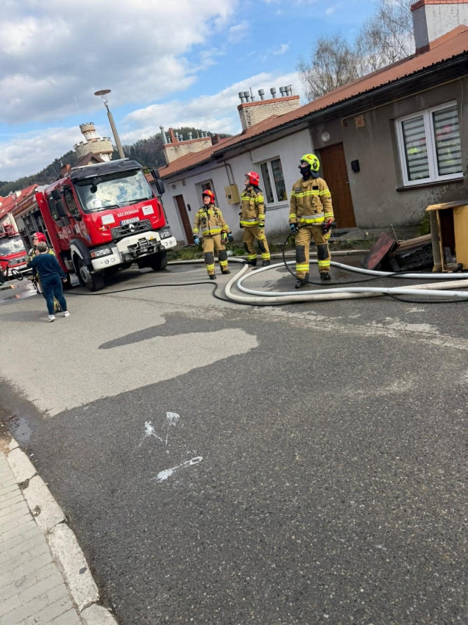 Firefighters in yellow turnout gear standing on a residential street with a red fire truck and coiled hoses nearby.